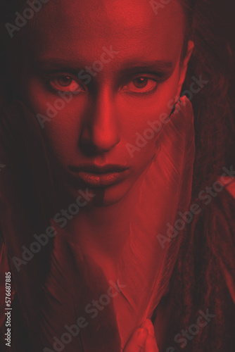 Beautiful woman studio portrait. Model with make-up and dreadlocks holding big bird feathers near her face and looking to camera with serious or seductive look. Toned image with red and black color