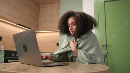 Mixed Race Woman Wrapped in Blanket Working Online. Female feeling cold working online from home office. Lack of heating in apartments.