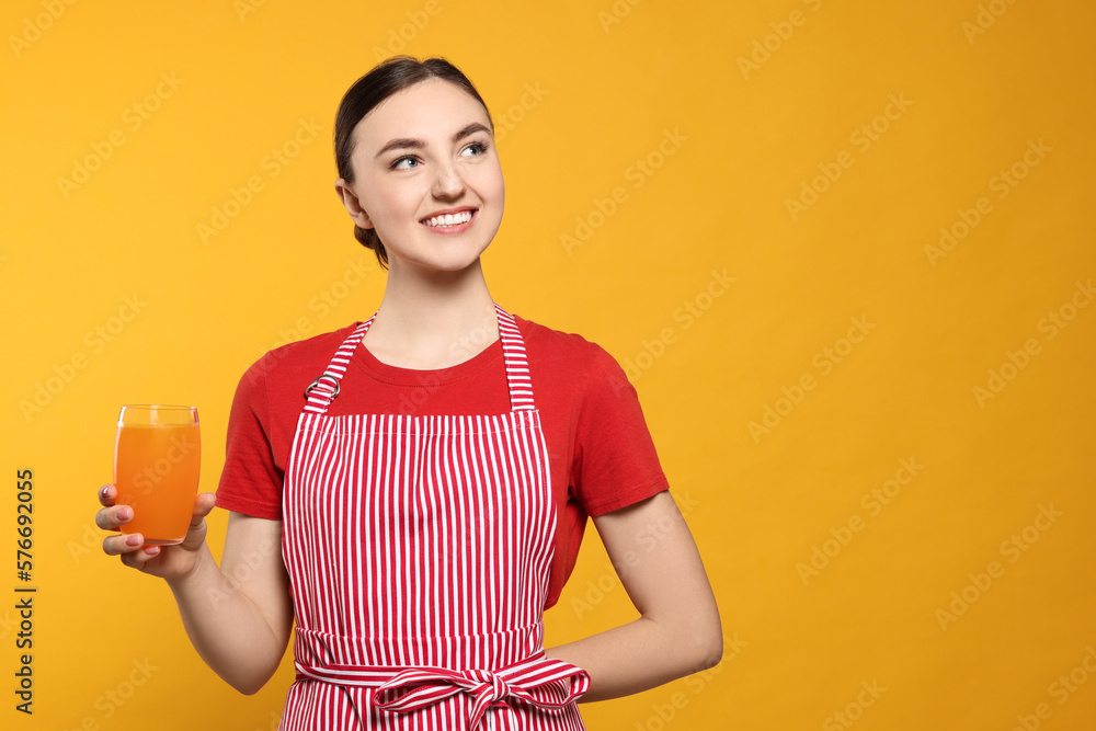 Beautiful young woman in clean striped apron with glass of juice on orange background. Space for text Beautiful young woman in clean striped apron with glass of juice on orange background. Space for text