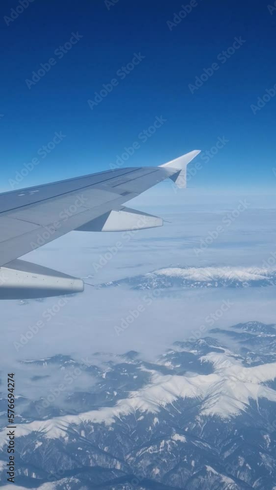 Plane flight above the Carpathian Mountains snowy peaks. Blue skyline and airplane wing seen through the window, vertical orientation Plane flight above the Carpathian Mountains snowy peaks. Blue skyline and airplane wing seen through the window, vertical orientation