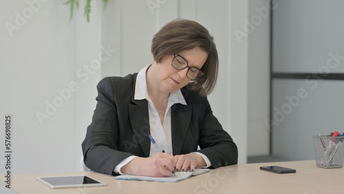 Old Businesswoman Writing while Sitting in Office © stockbakers