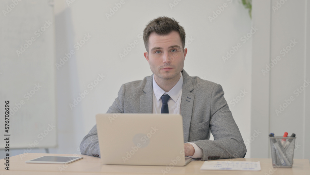 Young Businessman Looking at Camera while using Laptop Young Businessman Looking at Camera while using Laptop