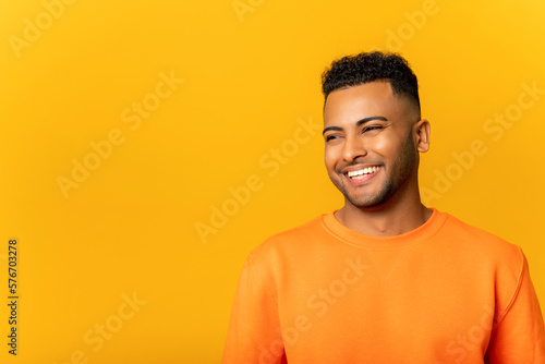 Portrait of happy satisfied handsome young Indian man standing with smile and looking away. Indoor studio shot on yellow background
