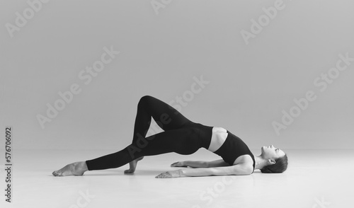 Keeping body fit and healthy. Black and white photography. Young girl doing stretching exercises over studio background. Concept of sport, body care, beauty, fitness, active lifestyle. Ad