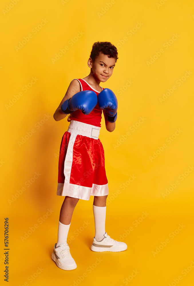 Active sportive little boy, beginner boxer in sports uniform and boxing gloves isolated over yellow background. Concept of sport, active and healthy lifestyle, kids emotions