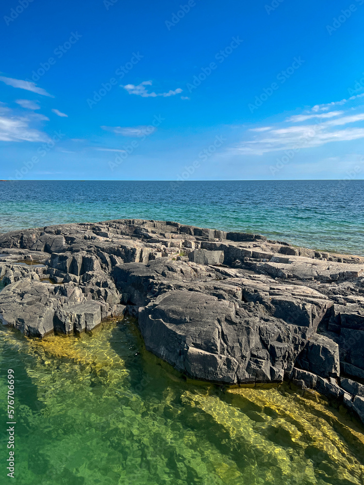 Large basin rock formation coast meets the crystal clear pure turquoise blue waters of Lake Superior. The rocks are jetting into the water. Daytime scene with blue skies above.