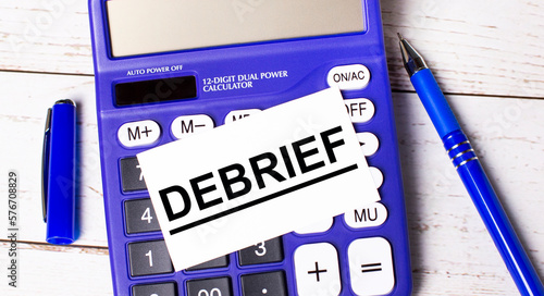 A large blue financial calculator, a blue pen and a blank card with the text DEBRIEF on a light wooden background. Concept of calculations, accounting, calculations, profit.