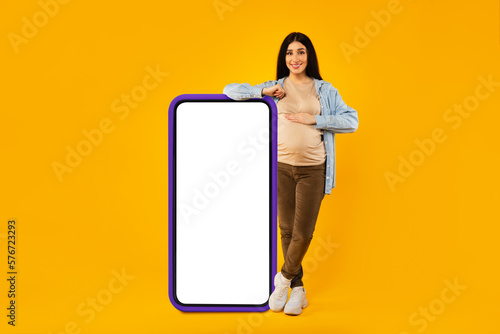 Pregnancy app. Happy expectant lady standing near huge smartphone with blank white screen, mockup