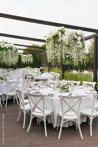 A general plan of a festive tables decorated with a white tablecloth, white chairs, plates, compositions of white flowers and candles. Hanging flower beds and light bulbs. Wedding decoration and decor