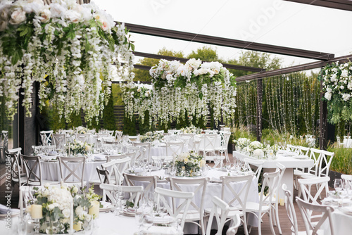 A general plan of a festive tables decorated with a white tablecloth, white chairs, plates, compositions of white flowers and candles. Hanging flower beds and light bulbs. Wedding decoration and decor