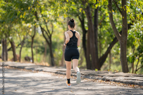 A young runner woman wearing fitness clothing runs in a city park surrounded by trees and greenery