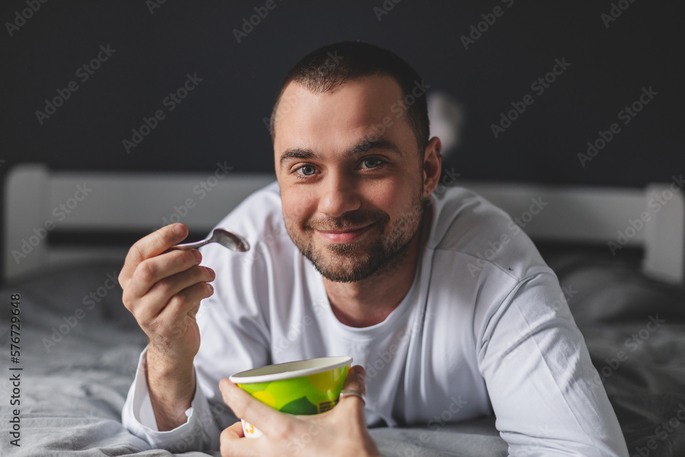 Handsome man lies on his bed while eating a pint of pistachio ice cream with spoon. He is look at window, dreaming. Eating in bed. Happy beautiful man resting in his comfortable bed at home. Yummy. Handsome man lies on his bed while eating a pint of pistachio ice cream with spoon. He is look at window, dreaming. Eating in bed. Happy beautiful man resting in his comfortable bed at home. Yummy.