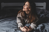 Young happy smile woman day dreaming think looking up hold cell phone on bed in bedroom, at home, girl lying type message, look at someone, flirting.