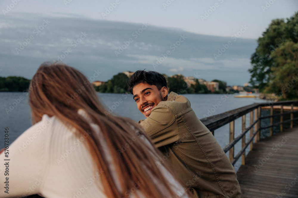 Side view of happy young man talking with female friend while leaning on railing at pier Side view of happy young man talking with female friend while leaning on railing at pier