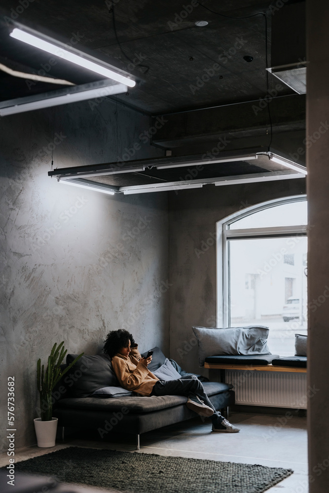 Female entrepreneur using mobile phone while sitting on sofa at creative workplace Female entrepreneur using mobile phone while sitting on sofa at creative workplace