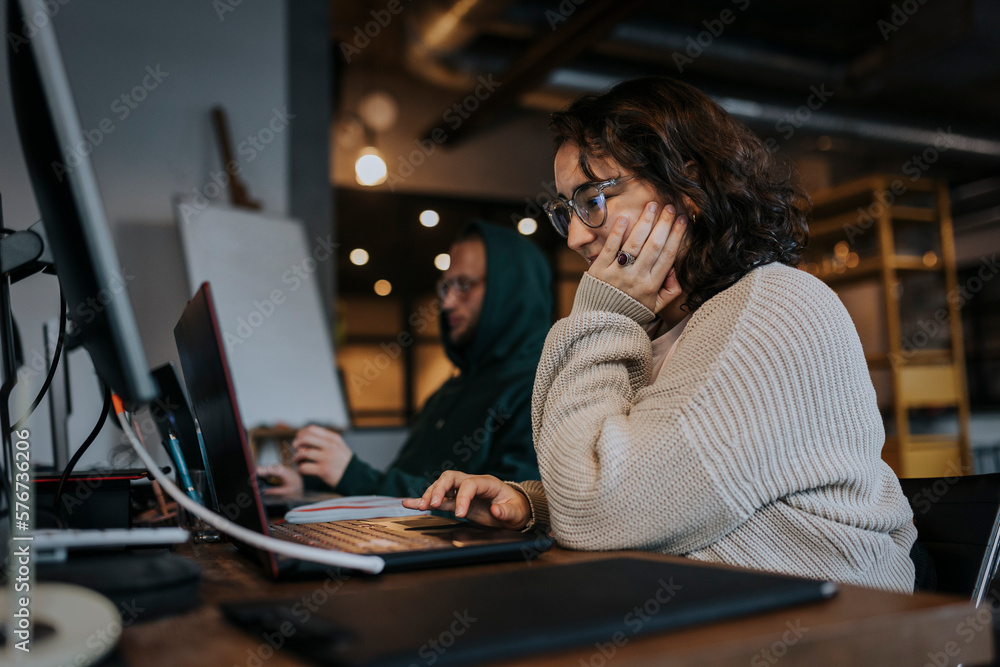 Young female hacker working on laptop by male colleague at desk in startup company Young female hacker working on laptop by male colleague at desk in startup company