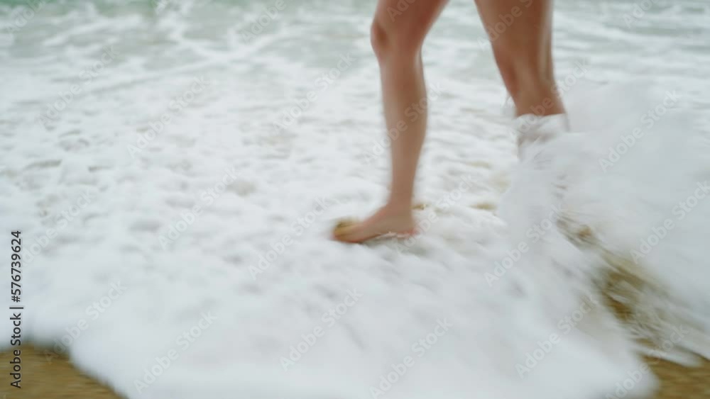Slim female legs and feet walking along sea water waves on sandy beach. Pretty woman walks at seaside surf. Splashes of water and foam. Girl legs walking along shore on sand in tide leaving footprints Slim female legs and feet walking along sea water waves on sandy beach. Pretty woman walks at seaside surf. Splashes of water and foam. Girl legs walking along shore on sand in tide leaving footprints