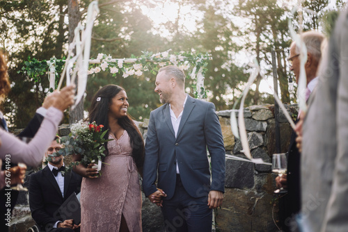 Cheerful newlywed couple holding hands while walking amidst guests at wedding ceremony photo