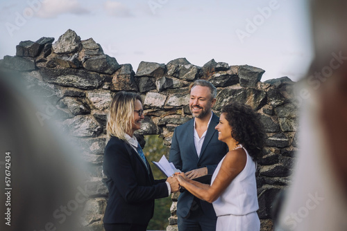Happy lesbian couple holding hands while minister looking at them against stone wall during wedding ceremony