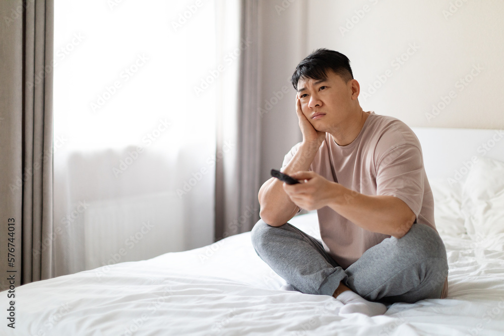 bored asian man sitting on bed at home, channels on tv bored asian man sitting on bed at home, channels on tv