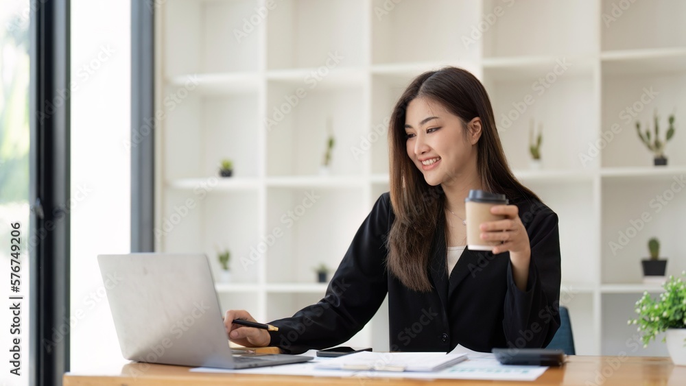 Asian business woman holding a cup of coffee at the desk Financial graph with desktop and laptop calculator for office marketing ideas Asian business woman holding a cup of coffee at the desk Financial graph with desktop and laptop calculator for office marketing ideas