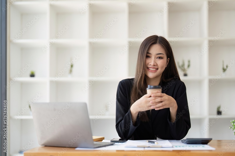 Asian business woman holding a cup of coffee at the desk Financial graph with desktop and laptop calculator for office marketing ideas Asian business woman holding a cup of coffee at the desk Financial graph with desktop and laptop calculator for office marketing ideas