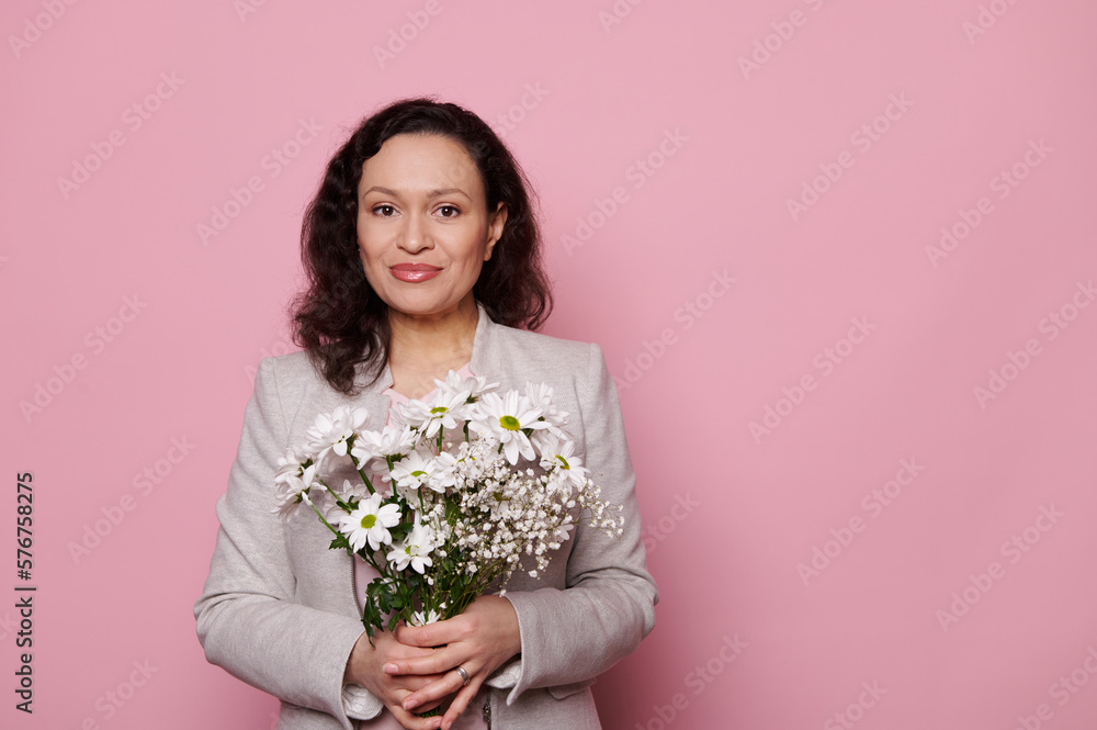 Beautiful multiracial middle-aged woman in stylish wear, holding a bouquet of spring flowers, smiling looking at camera,isolated on pink color background. Women's and Mother's Day concept. Copy space