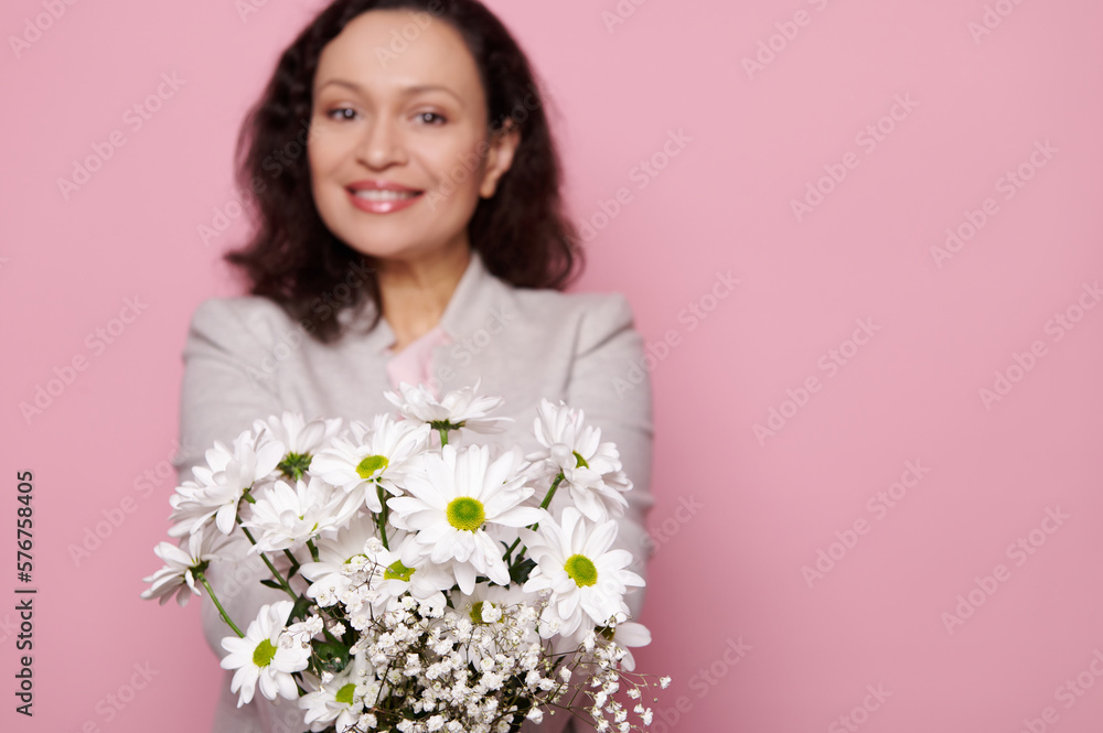 Cute spring bouquet of white chrysanthemums and gypsophila flowers, in hands of a blurred charming multi-ethnic woman, smiling a toothy smile looking at camera, isolated pink background. Copy ad space Cute spring bouquet of white chrysanthemums and gypsophila flowers, in hands of a blurred charming multi-ethnic woman, smiling a toothy smile looking at camera, isolated pink background. Copy ad space