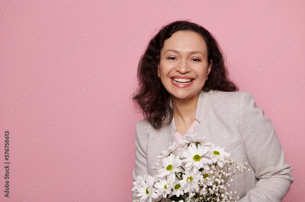 Happy mature woman with natural make-up, holding a bouquet of spring flowers and smiling a beautiful toothy smile looking at camera, isolated pink background. International Women's Day. Copy ad space Happy mature woman with natural make-up, holding a bouquet of spring flowers and smiling a beautiful toothy smile looking at camera, isolated pink background. International Women's Day. Copy ad space