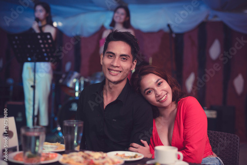 A sweet Asian couple smiles for the camera with the girlfriend resting her head on her boyfriend s shoulder as they dine in a restobar for a dinner date.