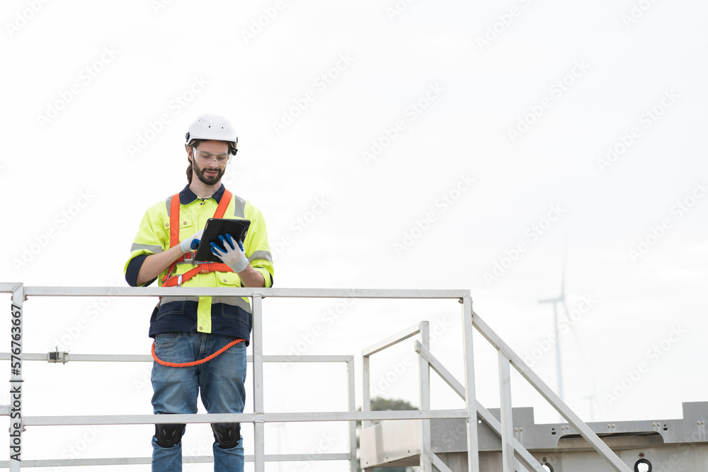 Male engineer wearing uniform and helmet while control or monitoring wind turbine by tablet at wind turbines farm Male engineer wearing uniform and helmet while control or monitoring wind turbine by tablet at wind turbines farm