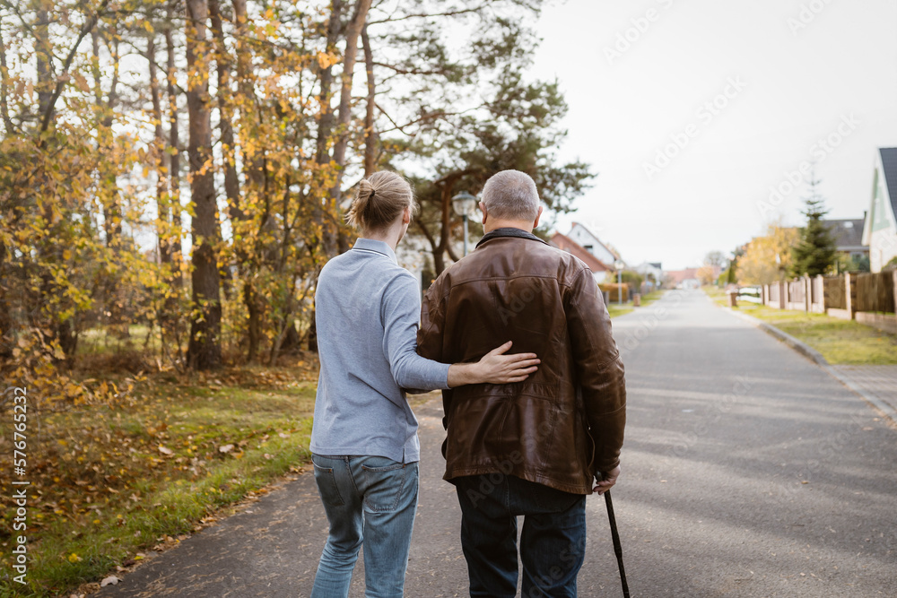 Rear view of caretaker supporting retired senior man walking with cane on road Rear view of caretaker supporting retired senior man walking with cane on road