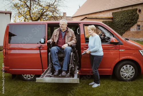 Smiling senior woman holding remote control while helping man with disability in motorized wheelchair to disembark from