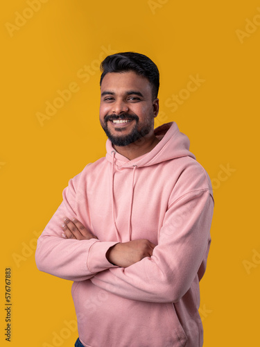 Smiling handsome Pakistani guy looking at camera. Casual pink hoodie crossed arms. Attractive toothy smile. Yellow background. Positive mood  © Iryna&Maya