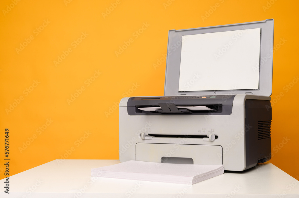 Front view of a multifunctional device -printer,copier,scanner and a stack of blank sheets of A4 paper on a white table on a yellow background, mockup.Office equipment, close-up.Multifunction printer. Front view of a multifunctional device -printer,copier,scanner and a stack of blank sheets of A4 paper on a white table on a yellow background, mockup.Office equipment, close-up.Multifunction printer.