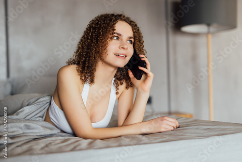 Undressed young woman lying in bed and talking on phone