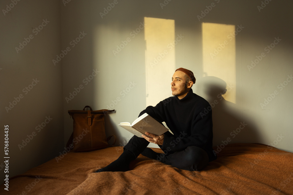 A caucasian man sitting on a bed lit by a window holding a book and wearing a knitted sweater and a beanie.