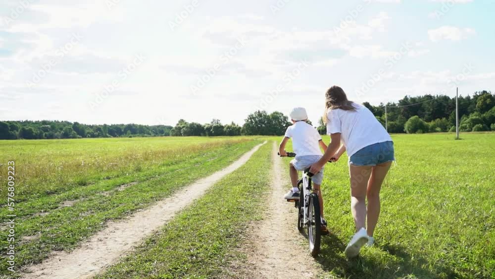 Childhood bike concept. Mother teaching son to ride bicycle. Happy cute boy learn to riding a bike in park at sunset time. Young mom teaching son to ride bike first time on countryside rural road.