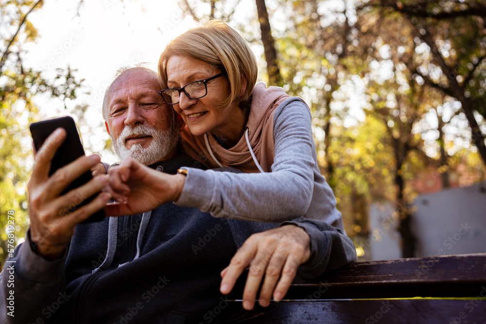 Cheerful seniors in sports clothing sitting on a park bench after exercise looking at smartphone, talking, smiling Cheerful seniors in sports clothing sitting on a park bench after exercise looking at smartphone, talking, smiling