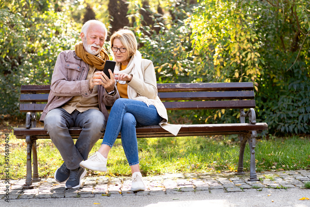 Cheerful elderly couple using banking apps on a smartphone to make payments outdoors in the park Cheerful elderly couple using banking apps on a smartphone to make payments outdoors in the park