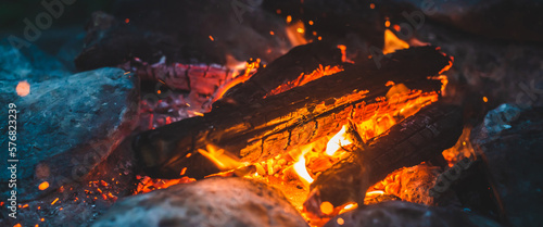 Vivid smoldered firewoods burned in fire close-up. Atmospheric background with orange flame of campfire. Unimaginable full frame image of bonfire. Glowing embers in air. Warm logs, bright sparks bokeh