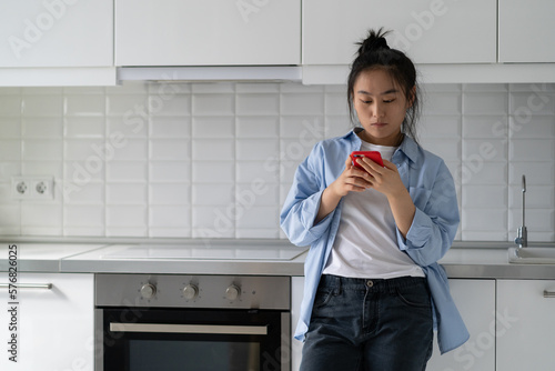 Surfing social media addicted Asian Korean woman stand at home puts things off until later, not concentrate and look away from mobile phone. Chinese girl use mobile phone in kitchen read online recipe