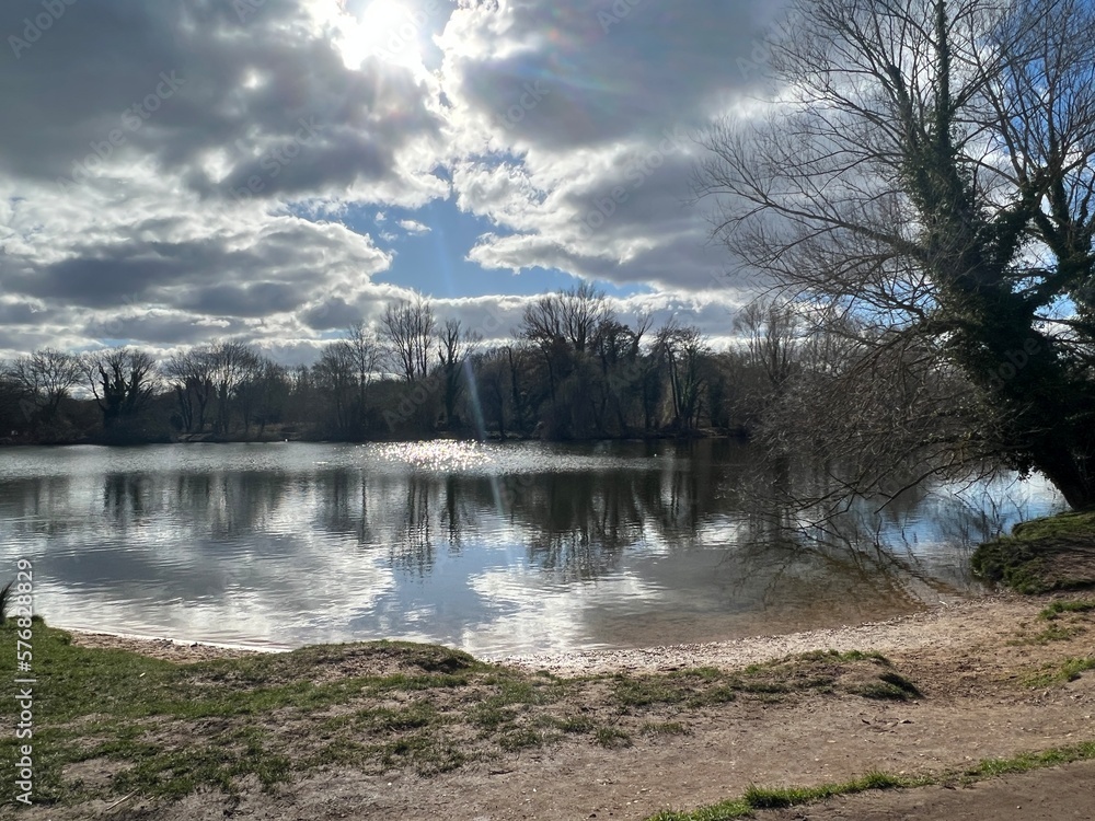 Fototapeta premium Landscape of Norfolk Broad with sandy shore by large lake , the Spring blue sky with sun light through white puffy cloud and trees reflected in calm river water on bright cold day in East Anglia UK