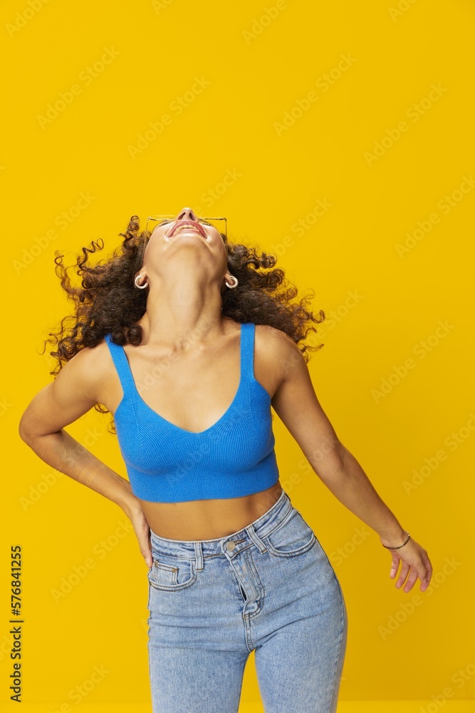 Woman with curly afro hair in a blue T-shirt on a yellow background dancing flying hair with sunglasses yellow, hand signs, look into the camera, smile with teeth and happiness, copy space Woman with curly afro hair in a blue T-shirt on a yellow background dancing flying hair with sunglasses yellow, hand signs, look into the camera, smile with teeth and happiness, copy space