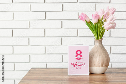 Vase with tulips and greeting card for Women's Day on table near white brick wall