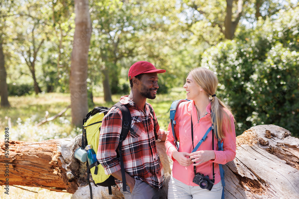 Happy diverse couple with backpacks trekking in sunny forest, smiling at each other Happy diverse couple with backpacks trekking in sunny forest, smiling at each other