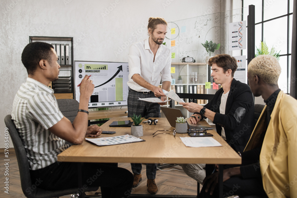 Fototapeta premium Multi ethnic male financiers discussing strategy solution for company development while sitting at office room. Group of four colleagues working with papers and modern gadgets.
