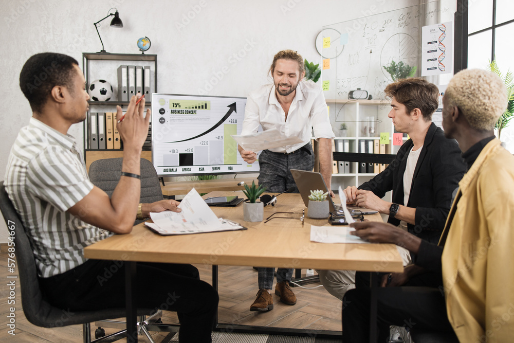 Caucasian businessman in white shirt smiling and showing financial report during conference with multiracial partners. Business people sitting at desk office with papers and modern technology. Caucasian businessman in white shirt smiling and showing financial report during conference with multiracial partners. Business people sitting at desk office with papers and modern technology.