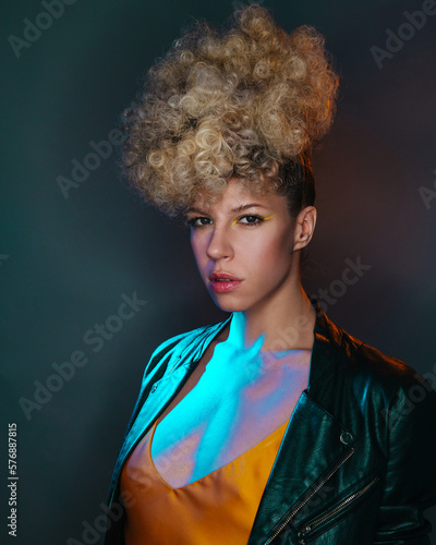 Curly dancer in night club. Fashion female model in color light on dark background. Close up of stylish girl in leather jacket.