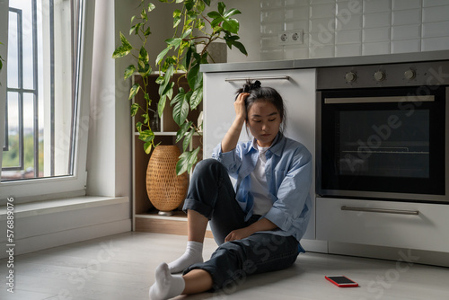 Upset depressed Asian girl suffering from breakup, heartbroken frustrated woman sits on floor in kitchen looking at smartphone waiting for call or message from ex boyfriend. Codependent relationships